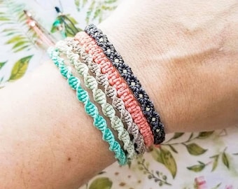 May include: A collection of five macrame bracelets in various colors: teal, light green, tan, coral, and dark gray. The bracelets are intricately woven with a spiral design, showcasing a handmade aesthetic. The bracelets are displayed on a wrist against a floral background.