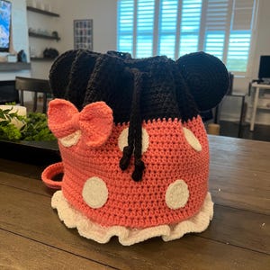 May include: A handmade crochet bag in the shape of a cartoon character's head. The bag is primarily coral pink with white polka dots and a white ruffled trim. It features black ears, a coral pink bow, and a drawstring closure.