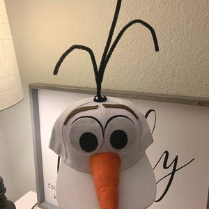May include: A white baseball cap with a felt snowman face, black eyes, a brown felt mouth, and an orange felt nose. The cap has black pipe cleaner arms attached to the top.