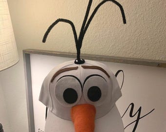 May include: A white baseball cap with a felt snowman face, black eyes, a brown felt mouth, and an orange felt nose. The cap has black pipe cleaner arms attached to the top.