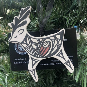 May include: A ceramic deer ornament with black and brown geometric designs. The deer is white with black antlers and a black ribbon for hanging. The ornament is hanging on a green Christmas tree.