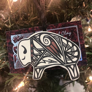 May include: A white ceramic ornament shaped like a buffalo with black and brown geometric patterns. The ornament is hanging on a Christmas tree with lights.
