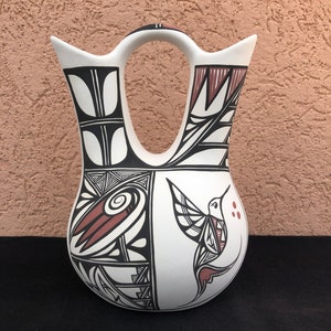 Peut inclure: Un vase en céramique blanc avec un design unique à double bec et une anse. Le vase est orné de motifs géométriques noirs et brun rougeâtre et d'une illustration de colibri. Le vase est probablement fait à la main et adapté à la décoration intérieure.
