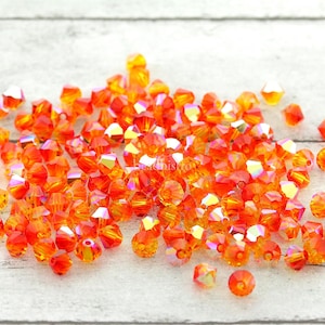 May include: A collection of orange and yellow faceted beads with an iridescent sheen. The beads are scattered on a light-coloured wooden surface. The beads are a mix of shapes and sizes, reflecting light.