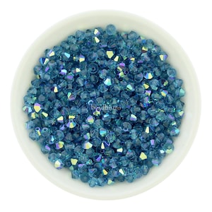 Puede incluir: Un cuenco blanco lleno de numerosas cuentas azules iridiscentes facetadas. Las cuentas tienen un aspecto cristalino, reflejando múltiples colores. La imagen está tomada desde una perspectiva cenital, mostrando la textura y las variaciones de color de las cuentas.