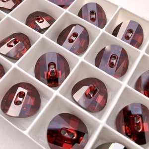May include: A close-up of a package of 18 red crystal buttons. The buttons are arranged in a grid pattern in a white plastic container. Each button has two holes and a faceted design.
