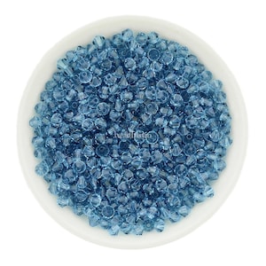 Puede incluir: Un cuenco de cerámica blanco lleno de numerosas cuentas de cristal azul claro. Las cuentas tienen una forma facetada en forma de cono, que refleja la luz. Las cuentas están dispersas por todo el cuenco, creando una superficie texturizada. La imagen está bien iluminada.