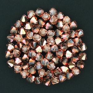 May include: A group of 50 rose gold crystal bicone beads. The beads are faceted and have a shiny finish.