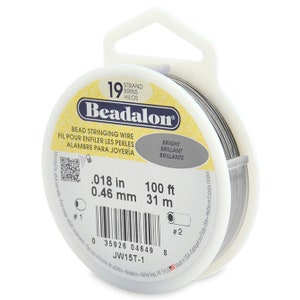 May include: A spool of 19 strand Beadalon bead stringing wire in a clear plastic container with a white label. The label says "Beadalon", "Bead Stringing Wire", "Fil Pour Enfiler Les Perles", "Alambre Para Joyeria", "Bright", "Brilliant", "Brillante", ".018 in", "0.46 mm", "100 ft", "31 m", "JW15T-1", and has a barcode.