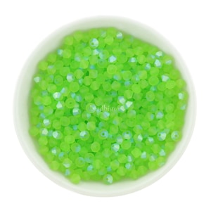 Op de afbeelding: Een kom gevuld met kleine, lichtgroene glazen kralen. De kralen hebben een gefacetteerde, iriserende afwerking.