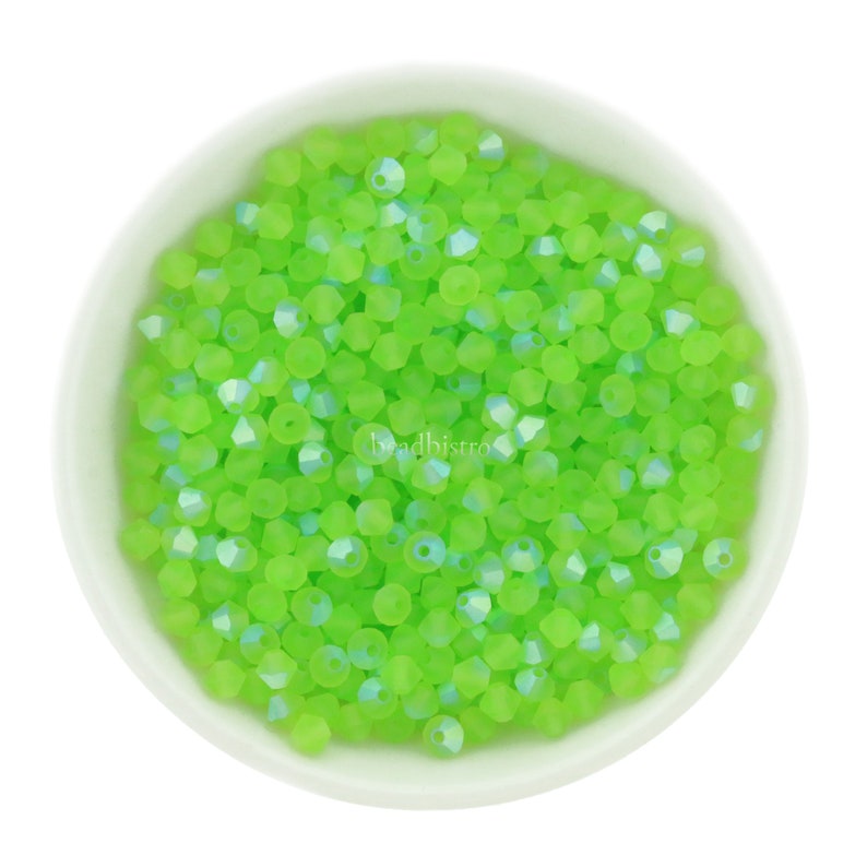 Op de afbeelding: Een kom gevuld met kleine, lichtgroene glazen kralen. De kralen hebben een gefacetteerde, iriserende afwerking.