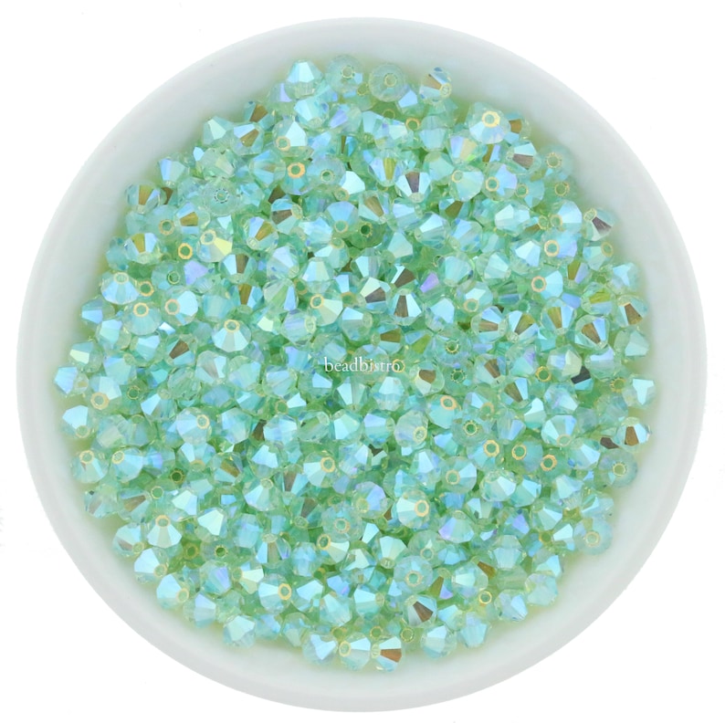May include: A bowl filled with light green glass beads. The beads are faceted and have a subtle iridescent sheen.