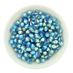 Puede incluir: Un cuenco blanco lleno de cuentas facetadas azules iridiscentes. Las cuentas tienen un diseño multifacético, que refleja la luz con una gama de tonos azules y amarillos. Las cuentas están dispersas por todo el cuenco, creando una exhibición visualmente atractiva.