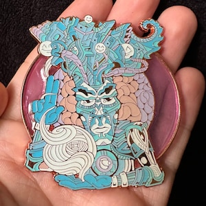 Puede incluir: Un pin de esmalte detallado con una figura estilizada en azul y turquesa, intrincados patrones geométricos y un fondo rosa. El pin tiene un borde color cobre e incluye varias caras sonrientes.