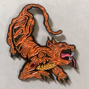 May include: Orange and black enamel pin depicting a snarling, stylized canine creature. The pin features intricate wood grain-like detailing, a pink tongue, and sharp white teeth. The creature is in a dynamic, stretched pose.