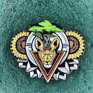 May include: A colorful enamel pin featuring a lion's face, sunflowers, and a tree with an hourglass. The pin has a geometric design with black, white, yellow, brown, green, and orange accents. The pin is approximately 2 inches in diameter.