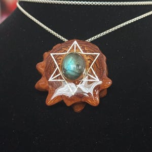 Puede incluir: Un collar con colgante con una base de madera marrón en forma de estrella y un diseño geométrico plateado. El colgante contiene una piedra preciosa azul-verdosa. La cadena del collar es plateada.