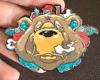 May include: A detailed enamel pin featuring a stylized bear with a city skyline on its head. The bear has a yellow face, brown fur, and is smoking a cigarette. The pin has blue, red, and white accents.