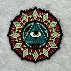 May include: A circular pin with a black border featuring a central teal triangle with an eye, surrounded by a geometric pattern in red, yellow, and cream colors. The pin has a detailed design and a glossy finish.