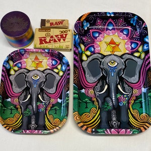 Puede incluir: Un conjunto de accesorios para fumar, que incluye dos bandejas metálicas con diseños de elefantes, un molinillo y dos paquetes de papel de liar RAW. Las bandejas presentan colores vibrantes y patrones intrincados.