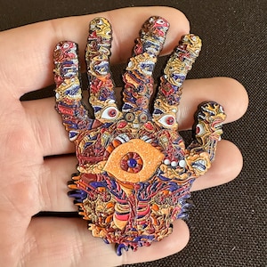 May include: A detailed, colorful pin in the shape of a hand, covered in intricate designs of eyes and faces. The pin features a central eye design and is predominantly orange, red, blue, and yellow. The pin is held in a hand.