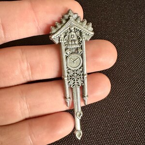 May include: A silver-colored miniature cuckoo clock pendant. The pendant features a detailed clock face, decorative scrollwork, and a roof-like structure at the top. The clock hands point to approximately 10:10. The pendant is held in a hand.