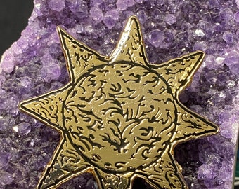 May include: A gold-colored, star-shaped pin with an intricate, swirling design in the center and along the edges. The pin is set against a backdrop of a purple amethyst crystal formation, creating a striking contrast.
