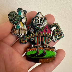 May include: A colorful enamel pin featuring a whimsical character holding a rooster and an egg. The pin has a black, red, and green color scheme with intricate details. The character stands on a patch of green grass.
