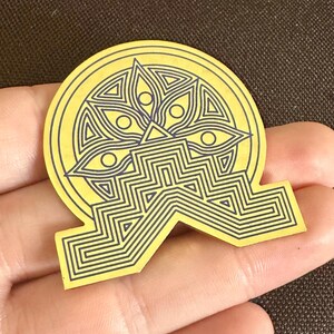 May include: A yellow sticker with a geometric design featuring a central eye motif and a series of interconnected lines. The sticker has a unique shape with a circular top and a stylized base.