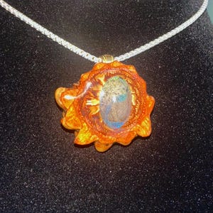 May include: A handcrafted pendant necklace featuring a unique, organic design. The pendant showcases a central, iridescent stone encased within a detailed, orange and yellow wooden frame. The necklace has a silver chain.