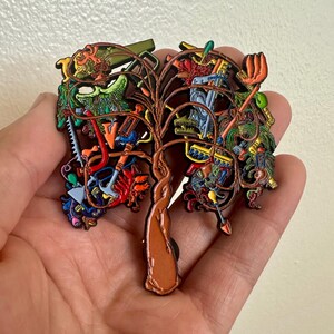 May include: A colorful enamel pin in the shape of a tree, featuring gardening tools as leaves. The pin has a brown trunk and branches, with a variety of tools in shades of red, green, blue, and orange.