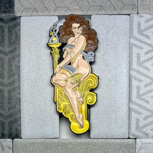 Puede incluir: Un pin que representa a una mujer desnuda con cabello castaño, sentada en un trono dorado ornamentado. El pin tiene un contorno negro e incluye una llama amarilla y detalles de humo. La mujer lleva una liga y una faja azul.