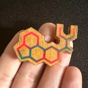 May include: Wooden pendant with a geometric design. The pendant features a series of connected hexagons, with red, teal, and purple accents. The pendant is held in a hand, and the background is a dark fabric.