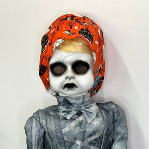 May include: A spooky doll with a cracked face, black eyes, and a gray dress. The doll wears a Halloween-themed orange head covering with black haunted house and tree designs. A gray bow tie and bow adorn the dress.