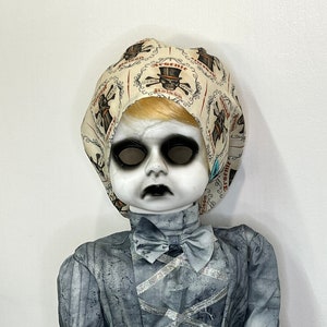 May include: A spooky doll with a pale face, black eyes and mouth, and blonde hair. The doll wears a gray dress with a bow tie and a patterned bonnet with the words "Arsenic Poison" and skull graphics. The dress has silver ribbon accents.