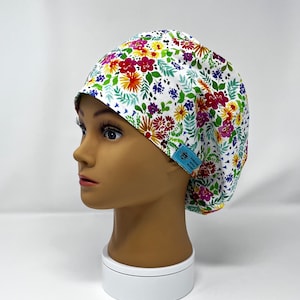 Puede incluir: Un gorro quirúrgico blanco con un vibrante estampado floral. El gorro presenta una colorida variedad de flores en tonos rojos, morados, amarillos y verdes. Una pequeña etiqueta azul con texto está adjunta al costado.