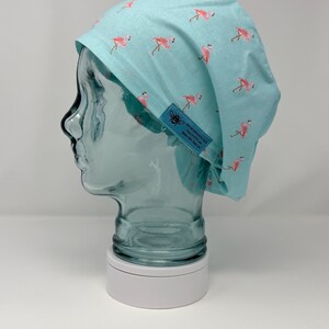 Puede incluir: Un gorro quirúrgico azul claro con un patrón repetido de flamencos rosas. El gorro tiene una pequeña etiqueta azul con un logotipo de abeja y texto. El gorro se muestra en una cabeza de maniquí transparente.