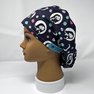 Puede incluir: Un gorro quirúrgico azul marino con un patrón repetido de un personaje de dibujos animados en una media luna, con lunares rosas, morados y turquesas. El gorro tiene una etiqueta azul con texto blanco. El gorro se muestra en una cabeza de maniquí.
