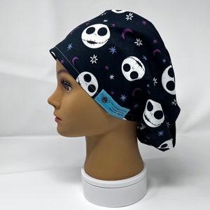 Puede incluir: Gorro quirúrgico negro con un patrón repetido de caras blancas de Jack Skellington, lunas crecientes moradas y estrellas. El gorro tiene una etiqueta azul claro con texto blanco. El gorro está sobre una cabeza de maniquí.