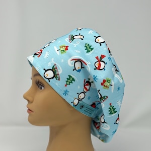 May include: A light blue scrub cap featuring a repeating pattern of penguins, snowflakes, and Christmas trees. The penguins are wearing scarves and hats. The cap is on a mannequin head.