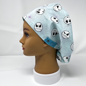 Puede incluir: Un gorro quirúrgico azul claro con un patrón repetido de caras de Jack Skellington de Pesadilla antes de Navidad. El gorro es blanco y negro con detalles en morado. Una pequeña etiqueta azul con texto está adjunta al costado.