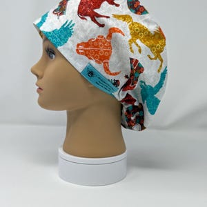 May include: A white scrub cap featuring colorful animal silhouettes in red, orange, yellow, and teal. The cap has a blue tag with text and a bee graphic. The cap is on a mannequin head.