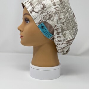 May include: A patterned scrub cap featuring a light brown and white design with botanical and postal-themed illustrations. The cap has a blue tag with a bee logo and text. The cap is displayed on a mannequin head.