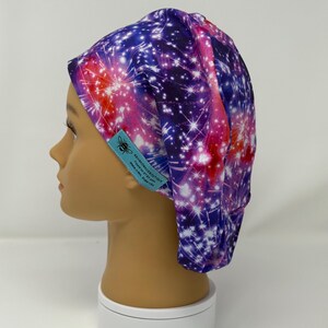 May include: A colorful scrub cap featuring a vibrant firework design in shades of purple, red, and white. The cap is made of fabric and has a tag with a bee logo and text. The cap is displayed on a mannequin head.