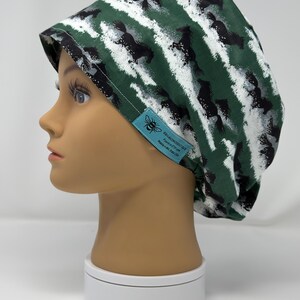 May include: A green and white patterned scrub cap featuring a repeating design of black horses. The cap has a tag with a bee logo and text. The cap is displayed on a mannequin head.