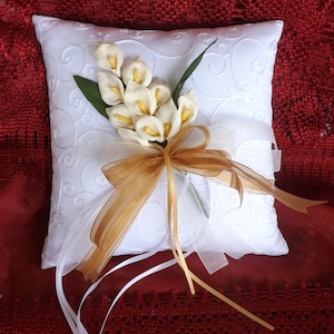 May include: White satin pillow with a gold ribbon and a bouquet of white calla lilies. The pillow is embroidered with a delicate floral pattern.