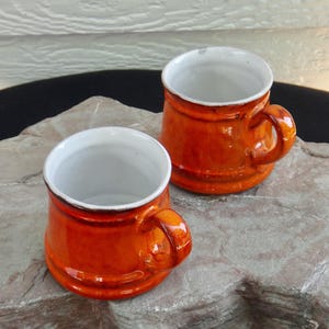May include: Two small, orange ceramic mugs with white interiors and rounded handles. The mugs have a glossy finish and sit on a stone surface. The mugs are likely for drinking beverages.