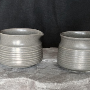 May include: A grey ceramic creamer with a handle and spout, and a matching sugar bowl. Both pieces feature horizontal ridged detailing around the body. The set is displayed on a stone-like surface against a dark background.
