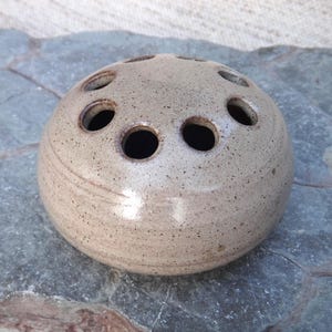 Peut inclure: Un petit vase rond en céramique beige avec plusieurs trous pour les tiges de fleurs. Le vase a un glaçage moucheté et une surface légèrement texturée.