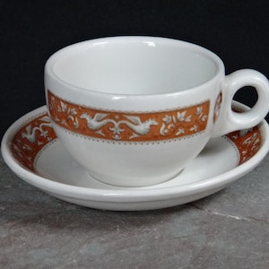May include: A white teacup and saucer with a brown and white floral pattern. The cup has a rounded handle and the saucer has a scalloped edge.